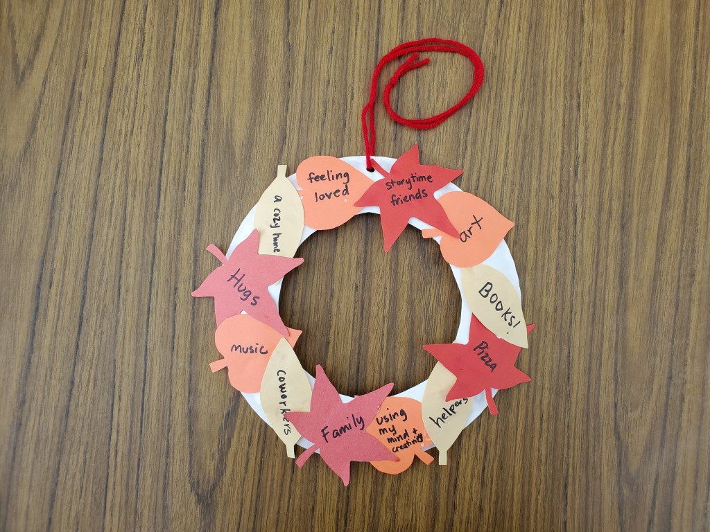 Photo of "thankful wreath" craft with orange, yellow, and red leaves with writing on them such as "books" "pizza" "family" "art" "storytime friends" etc.