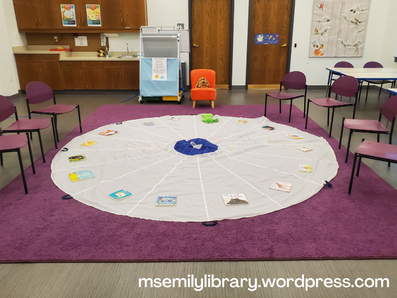 Photo showing Book Babies setup, with a round white parachute spread on a purple rug, with purple chairs to either side. A board book is set on each wedge, and a small pink chair sits at the head, with a dog puppet on the chair and a frog stuffy laying on the parachute. An easel to the side of the chair has song lyrics on it.