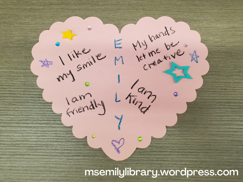 Craft showing a pink scalloped heart shape with the name EMILY written vertically down the middle, with "I like my smile" "My hands let me be creative" "I am friendly" "I am kind" also written on it, decorated with hearts, stars, stickers, and jewels.